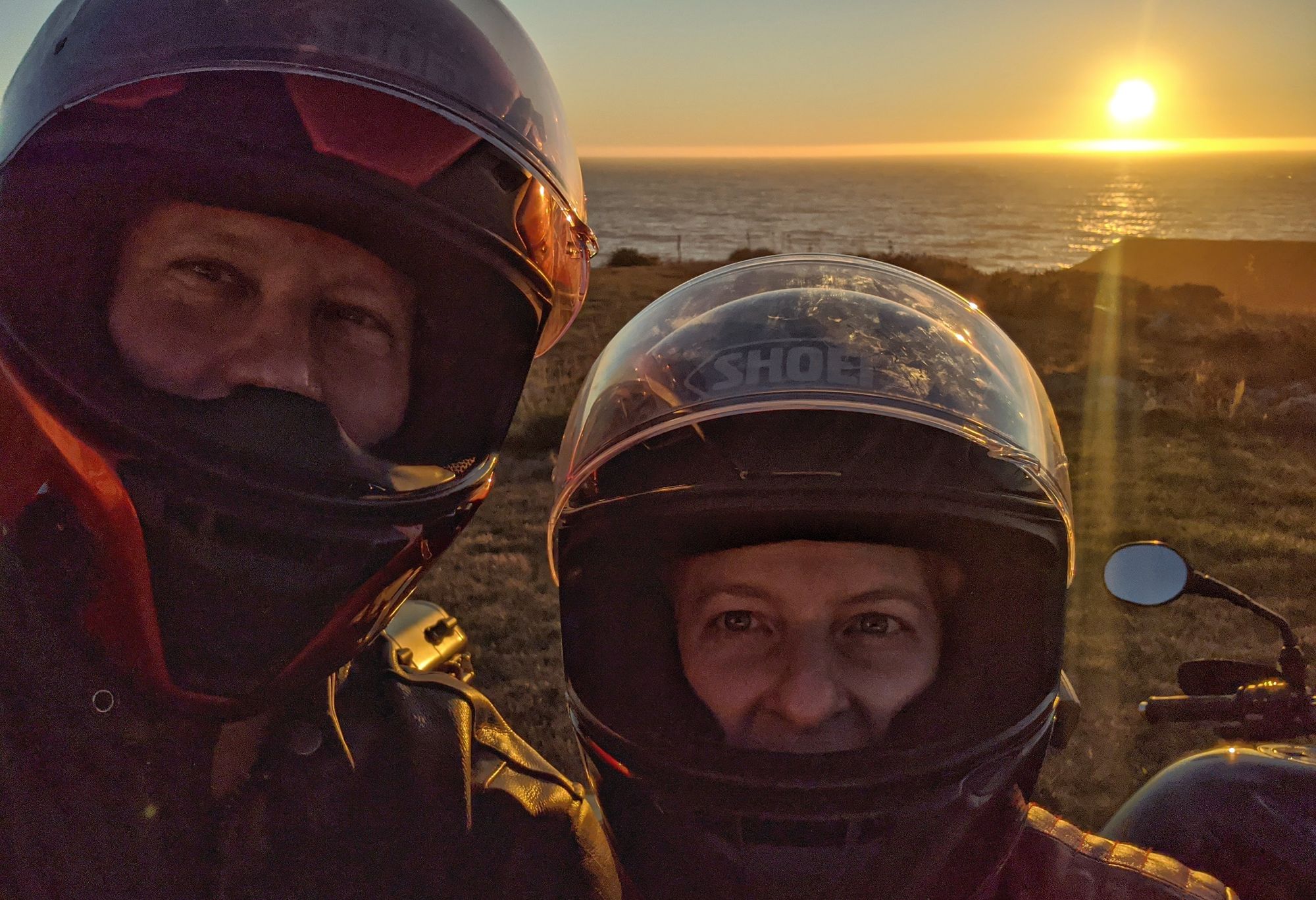 Seth and his wife wearing motorcycle helmets in front of a coastal sunset, northern California.